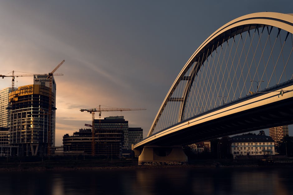 A skyline with modern bridge and construction cranes at sunset over a city river.